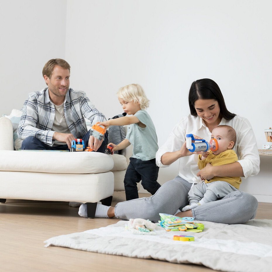 Young family sits in the living room, father plays with the toddler, mother sits with the baby on the floor and helps the baby to inhale asthma spray with a spacer
