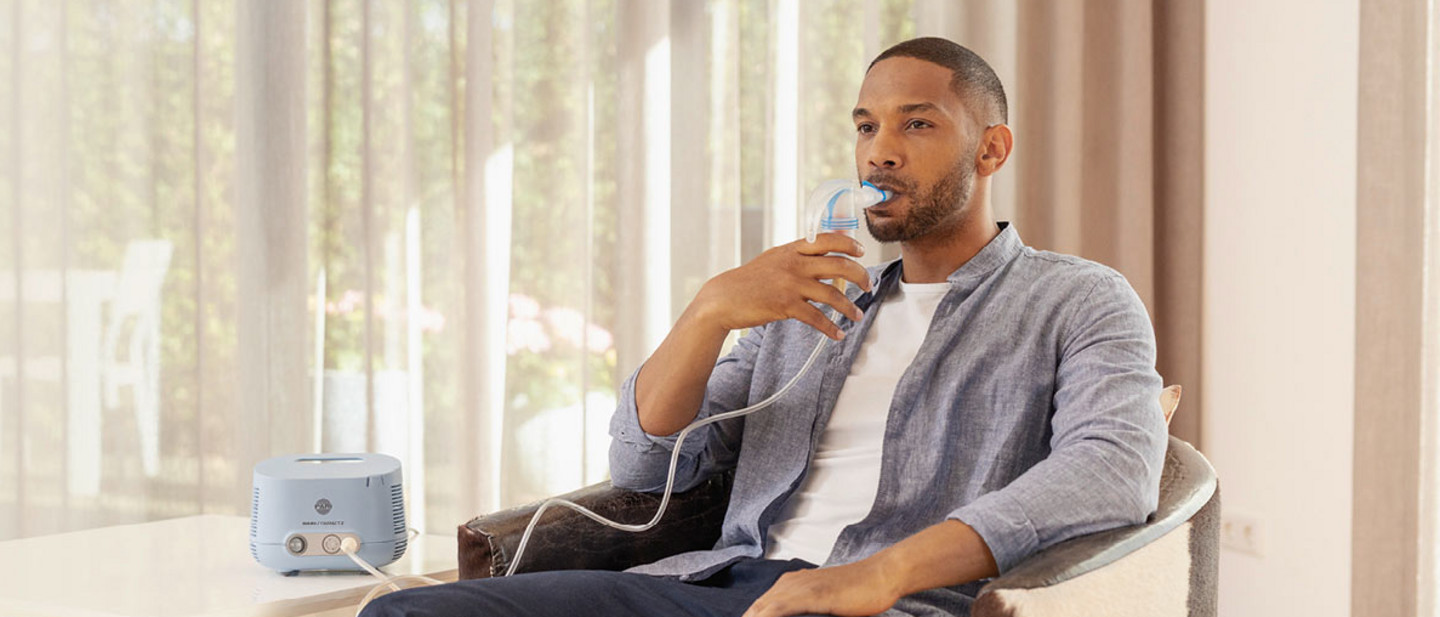 Ein Mann mit kurzem Bart und kurzen Haaren sitzt entspannt in einem Sessel und nutzt ein PARI COMPACT2 Inhalationsgerät. Er hält ein Mundstück mit einem Schlauch verbunden an den Mund, während das blaue Inhalationsgerät auf einem Tisch neben ihm steht. Durch ein großes Fenster im Hintergrund fällt Tageslicht in den Raum, leichte Vorhänge sind zu sehen.