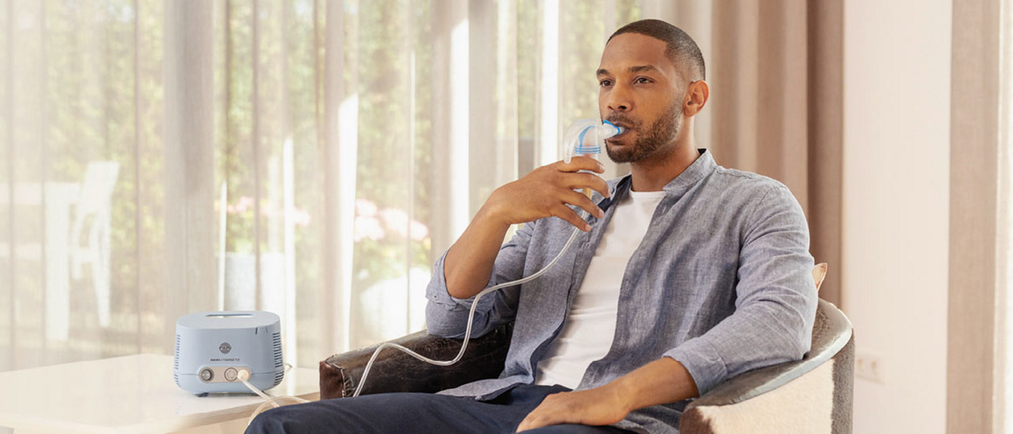 Ein Mann mit kurzem Bart und kurzen Haaren sitzt entspannt in einem Sessel und nutzt ein PARI COMPACT2 Inhalationsgerät. Er hält ein Mundstück mit einem Schlauch verbunden an den Mund, während das blaue Inhalationsgerät auf einem Tisch neben ihm steht. Durch ein großes Fenster im Hintergrund fällt Tageslicht in den Raum, leichte Vorhänge sind zu sehen.