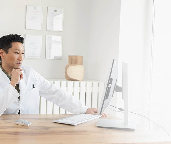 A doctor in a white coat is sitting at a desk, working intently on a computer in a bright office.