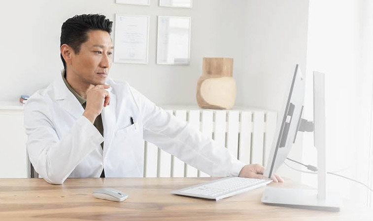 A doctor in a white coat is sitting at a desk, working intently on a computer in a bright office.