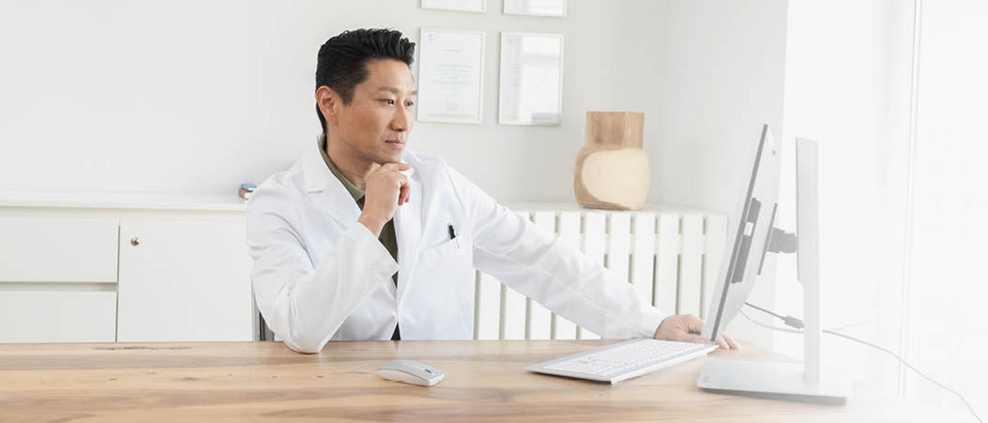 A doctor in a white coat is sitting at a desk, working intently on a computer in a bright office.
