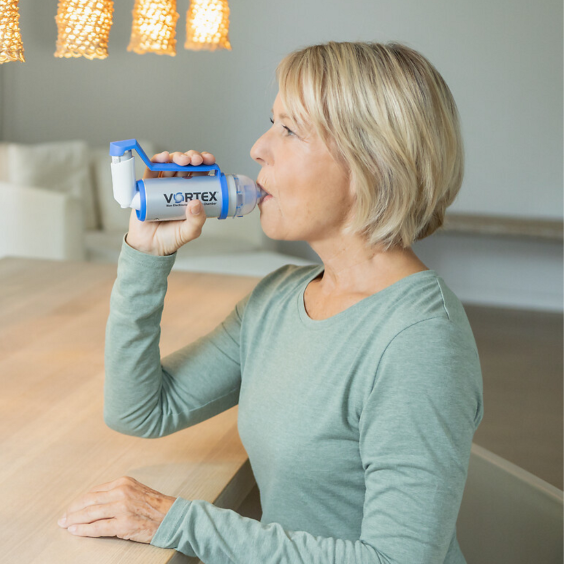 Ältere Frau sitzt im Esszimmer am Tisch und inhaliert ein Spray mit der Inhalierhilfe VORTEX