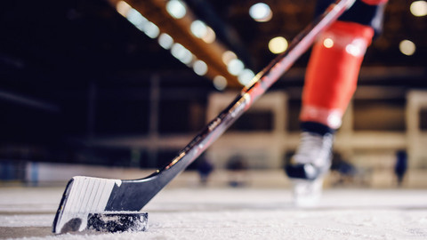 Nahaufnahme eines Hockeyschlägers mit Puk auf dem Eis, im Hintergrund sind Beine des Spielers zu erkennen