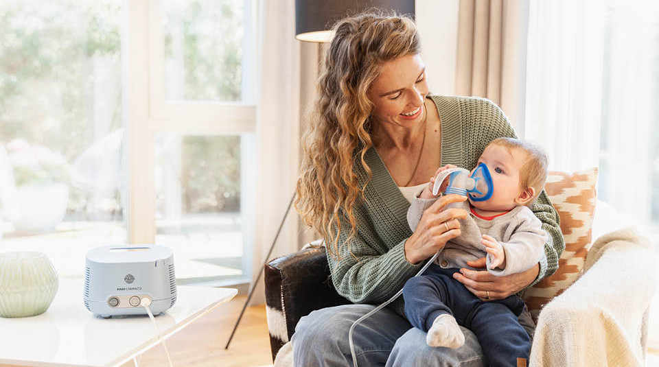 Erwachsene Person verabreicht einem Baby mit Atemmaske eine Inhalationstherapie mit einem PARI COMPACT Gerät im Wohnzimmer.