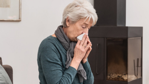 Ältere Frau sitzt auf der Couch im Wohnzimmer und putzt sich die Nase