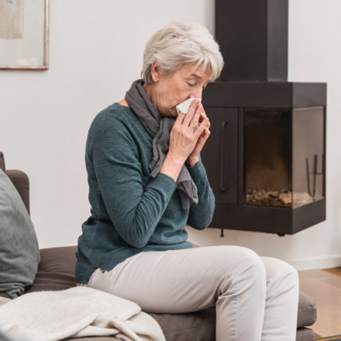 Ältere Frau sitzt auf der Couch im Wohnzimmer und putzt sich die Nase