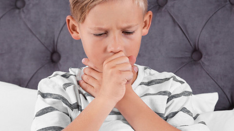 Junge sitzt in Bett und hustet in Faust, während er mit der anderen Hand um seinen Hals greift