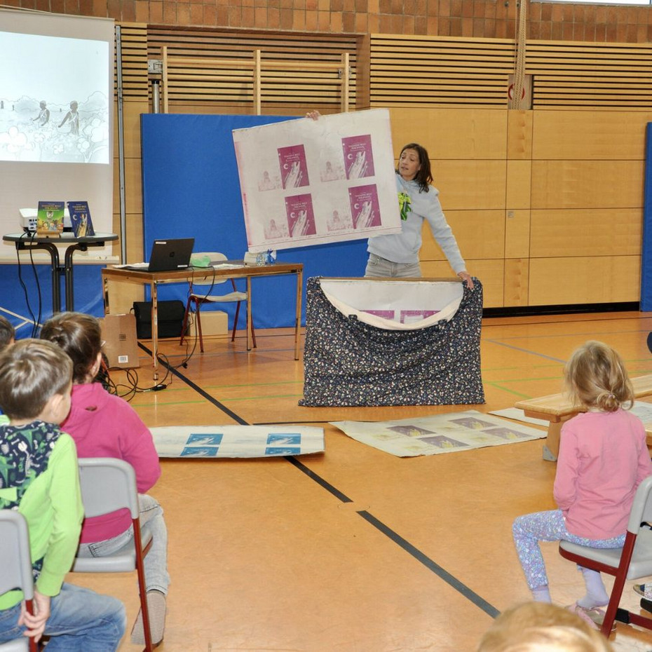 Author stands in front of seated children in the gym and holds up the printing plates of her book
