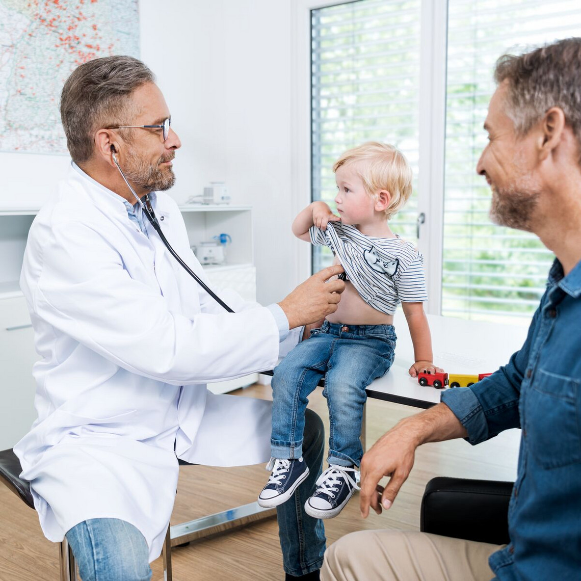 Paediatrician examines toddler with stethoscope, father sits next to him