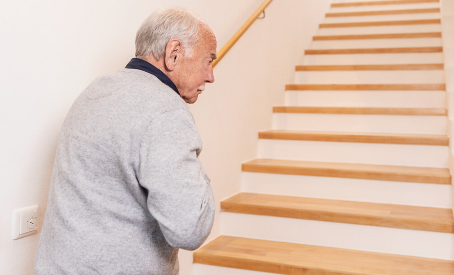 Älterer Mann steht an Fuß einer Treppe und hält sich die Brust