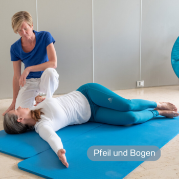 Aufnahme von der Seite: Junge Frau liegt auf einer Matte am Boden und macht die Übung Pfeil und Bogen, während die Physiotherapeutin sie anleitet