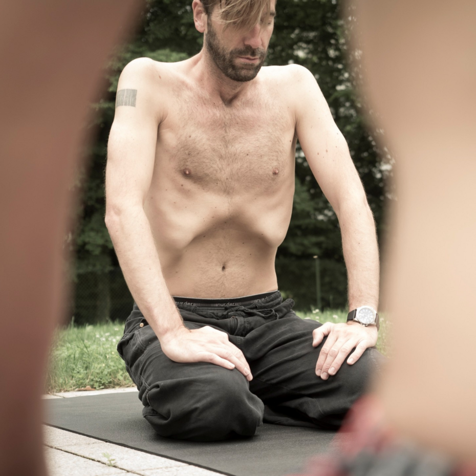 Mann sitzt auf seinen Knien am Boden, er konzentriert sich und atmet tief ein, Bauch nach innen gewölbt