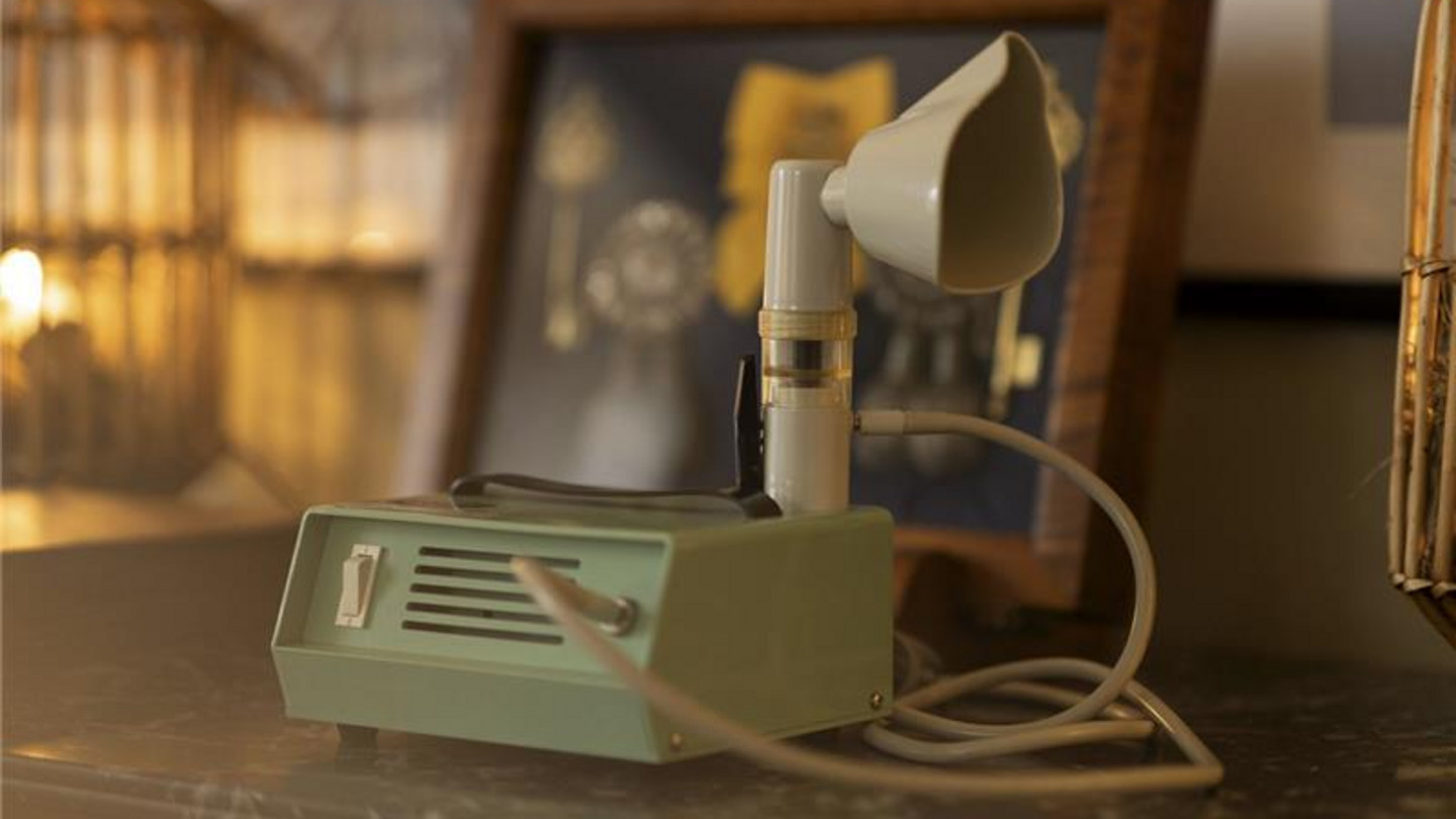 Vintage-Inhalationsgerät mit Vernebler Vintage-Inhalationsgerät mit Vernebler und Verbindungskabel auf einem Tisch, im Hintergrund ein gerahmtes Bild.