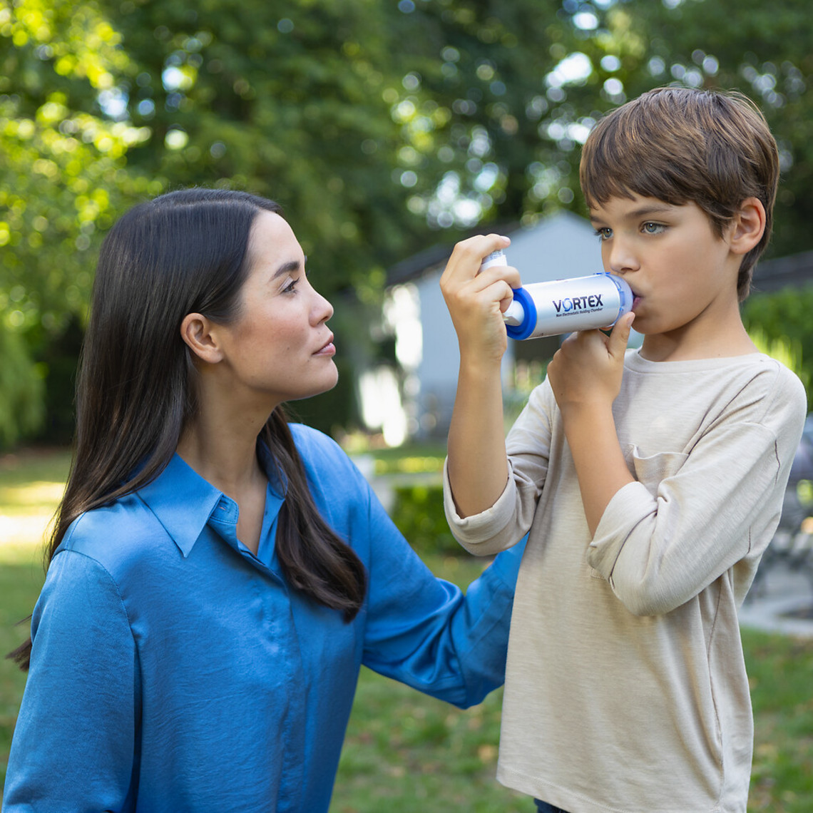 Mutter kniet vor Sohn im Garten, der Asthmaspray mit Inhalierhilfe VORTEX inhaliert
