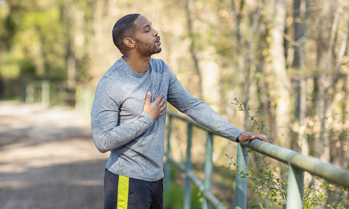 Eine Mann hält beim Joggen auf einem Waldweg an, stützt sich am Geländer ab und fasst sich mit der anderen Hand an die Brust.