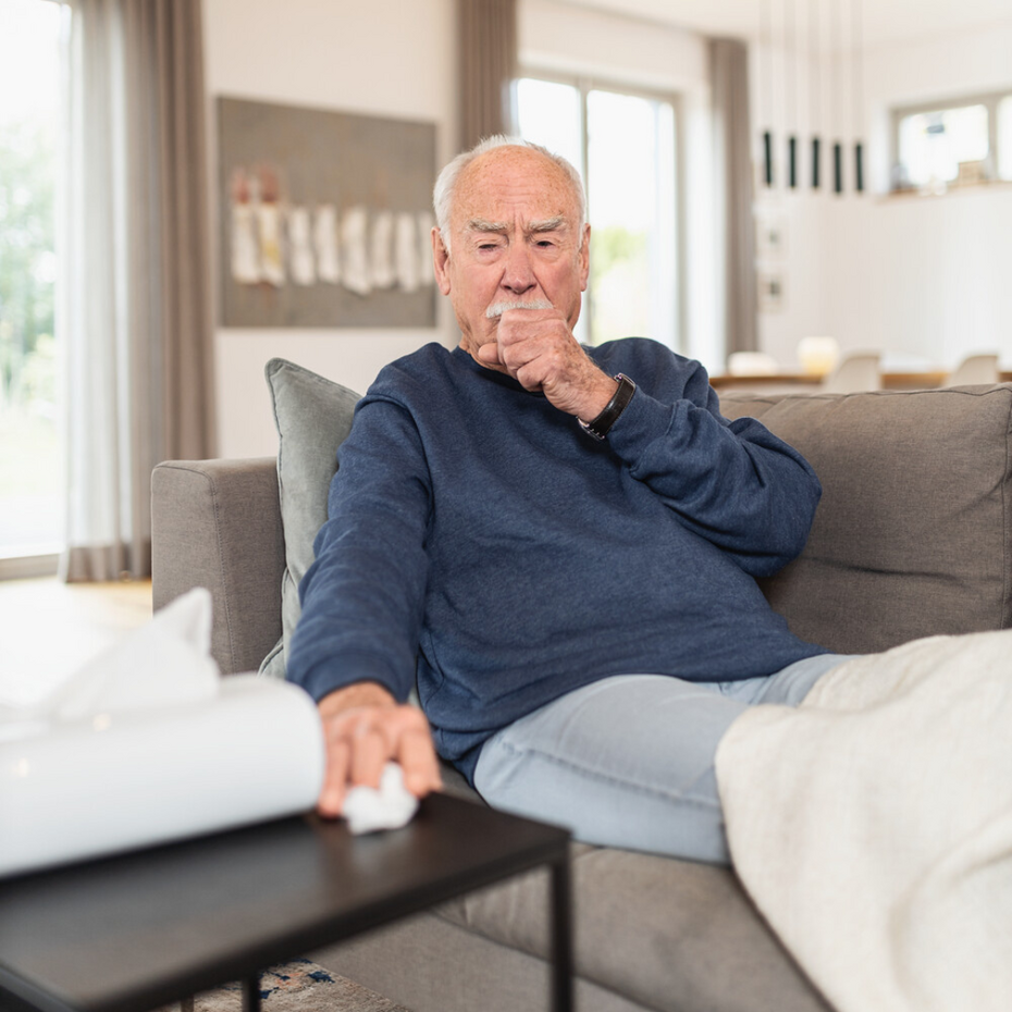 Älterer Mann sitzt auf der Couch und hustet in die geschlossene Faust