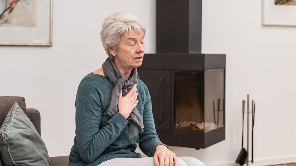 Ältere Frau atmet auf der Couch sitzend flach ein, um Hustenreiz zu unterdrücken