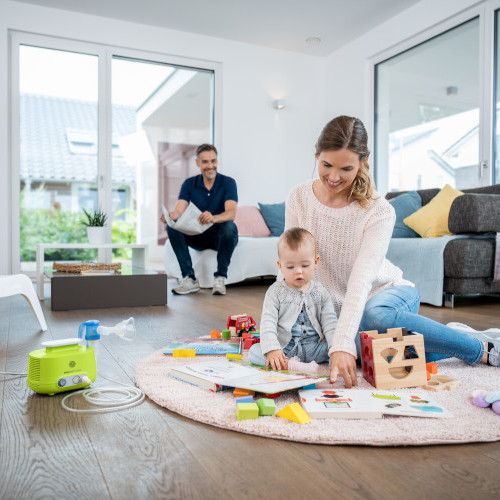 Hypertone Kochsalzlösung geeignet für Kinder und Babys Mutter sitzt mit Kleinkind auf dem Teppich und spielt, daneben steht ein Inhalationsgerät, Vater sitzt im Hintergrund auf Sofa