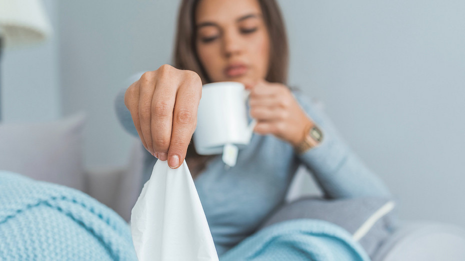 Woman sitting in bed under a blue blanket, holding a tissue and a white mug, appearing unwell or sick.