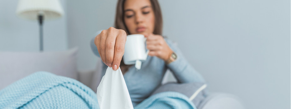 Woman sitting in bed under a blue blanket, holding a tissue and a white mug, appearing unwell or sick.