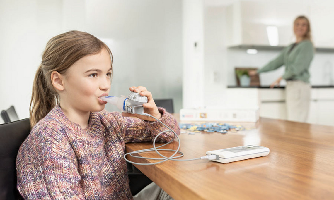 Mädchen sitzt am Holztisch und inhaliert mit einem mobilen Inhalationsgerät, während ihre Mutter im Hintergrund in der Küche steht.