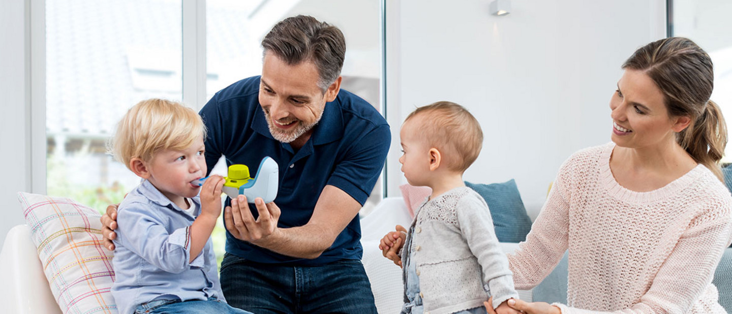 Ein Vater hilft seinem kleinen Jungen bei der Benutzung eines Inhalationsgeräts, während die Mutter die Hand eines zweiten jüngeren Kindes hält und lächelnd zusieht.