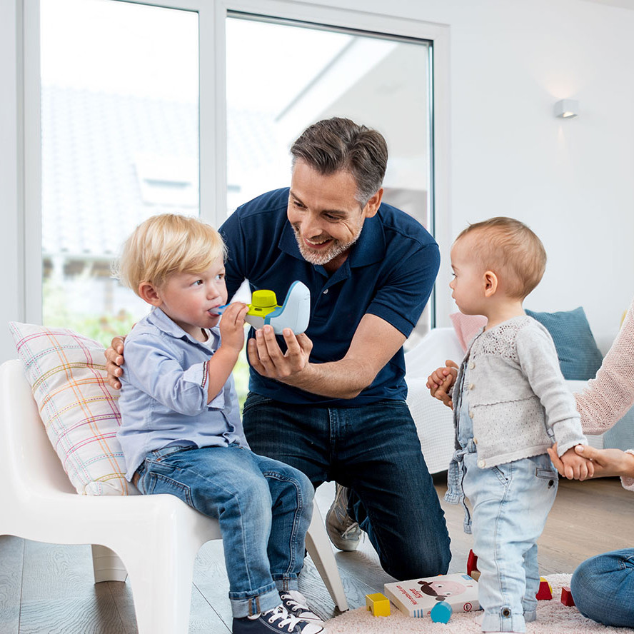 Ein Vater hilft seinem kleinen Jungen bei der Benutzung eines Inhalationsgeräts, während die Mutter die Hand eines zweiten jüngeren Kindes hält und lächelnd zusieht.