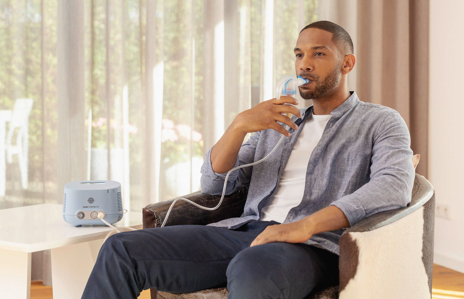 Ein Mann sitzt entspannt in einem Sessel und inhaliert mit dem PARI COMPACT2 Inhalationsgerät.