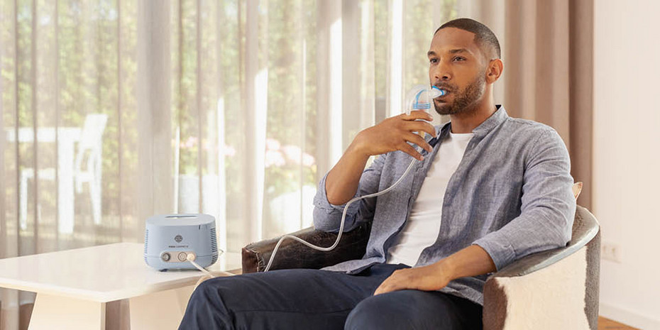 A young man inhales relaxed in a living room armchair using a PARI inhalation device