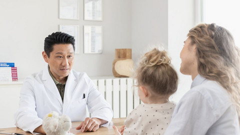 Arzt sitzt auf der einen Seite seines Tisches, er unterhält sich mit einer Mutter, die ihre Tochter auf dem Schoß hat, und auf der anderen Seite des Tisches sitzt