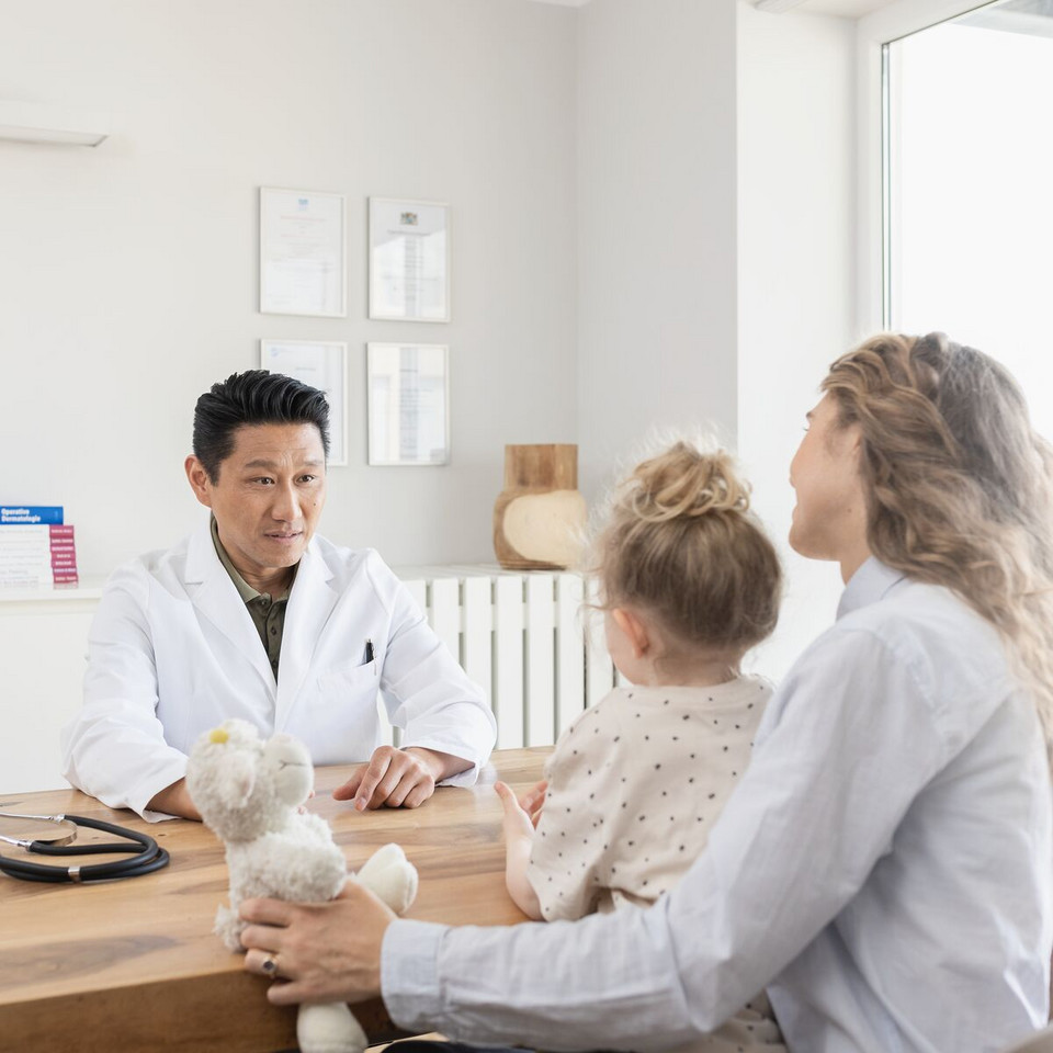 Calming irritated bronchial tubes: This helps manage bronchial symptoms Arzt sitzt auf der einen Seite seines Tisches, er unterhält sich mit einer Mutter, die ihre Tochter auf dem Schoß hat, und auf der anderen Seite des Tisches sitzt