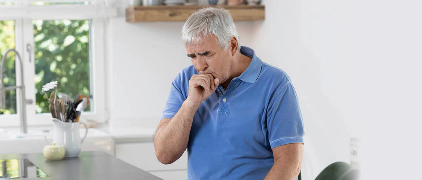 Bronchiektasen – krankhafte Erweiterungen der Bronchien Ein älterer Mann steht in einer Küche und hustet in seine Hand. Er stützt sich auf die Arbeitsplatte.