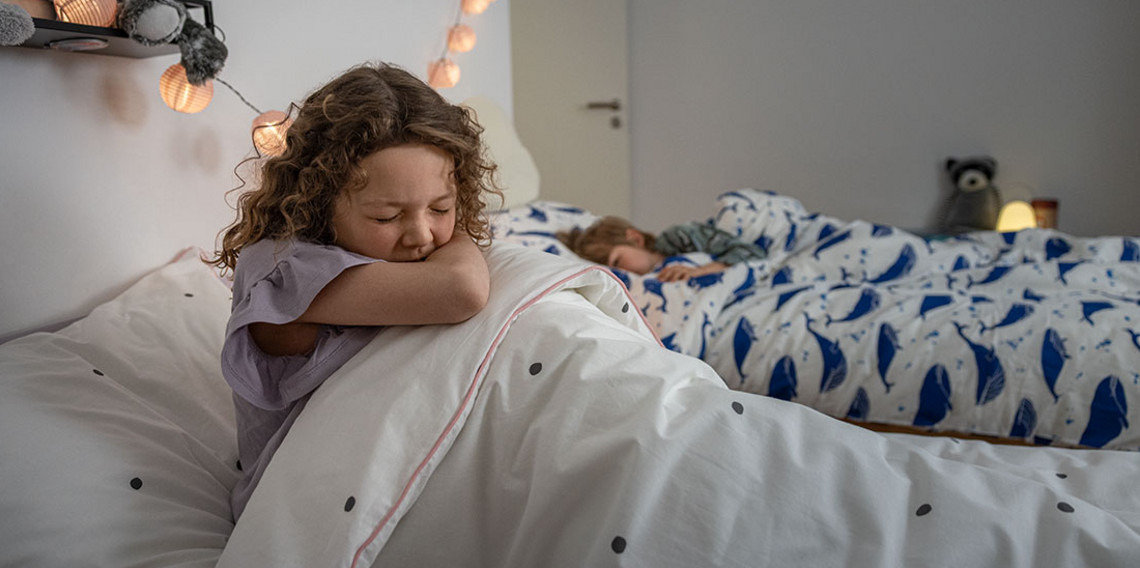 Kind sitzt im Bett und hustet in die Bettdecke Kind sitzt im Bett und hustet in die Bettdecke, während ein weiteres Kind im Hintergrund schläft.