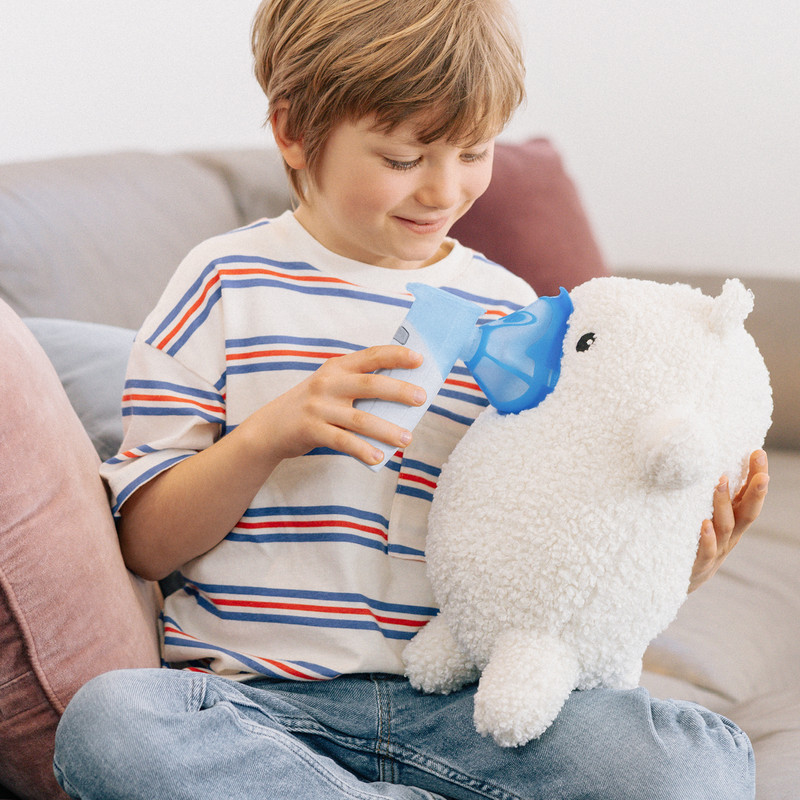 Kind sitzt auf einem Sofa und hält einem Kuscheltier eine Inhalationshilfe mit Maske vor das Gesicht.