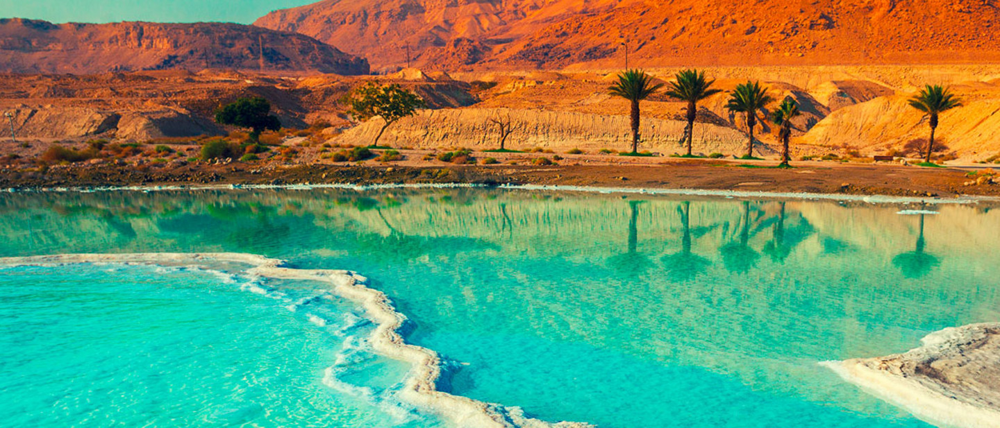 Eine beeindruckende Landschaft mit einer türkisblauen Salzlagune, umgeben von weißen Salzablagerungen. Im Hintergrund erheben sich rötlich-orange Wüstenberge, während eine Reihe von Palmen sich im ruhigen Wasser spiegelt.