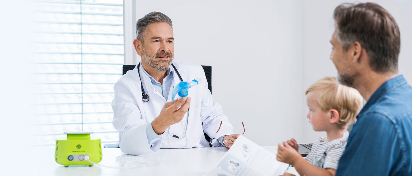 Vernebler Freundlich lächelnder Arzt in weißem Kittel sitzt an einem Schreibtisch mit einem PARI LC SPRINT Vernebler in der Hand. Ihm gegenüber sitzt eine Vater im blauen Jeanshemd. Er blättert in einer Anleitung und hat seinen kleinen blonden Sohn auf dem Schoß, der fasziniert den Vernebler begutachtet. Links neben dem Arzt steht ein grüner PARI BOY Junior Kompressor.