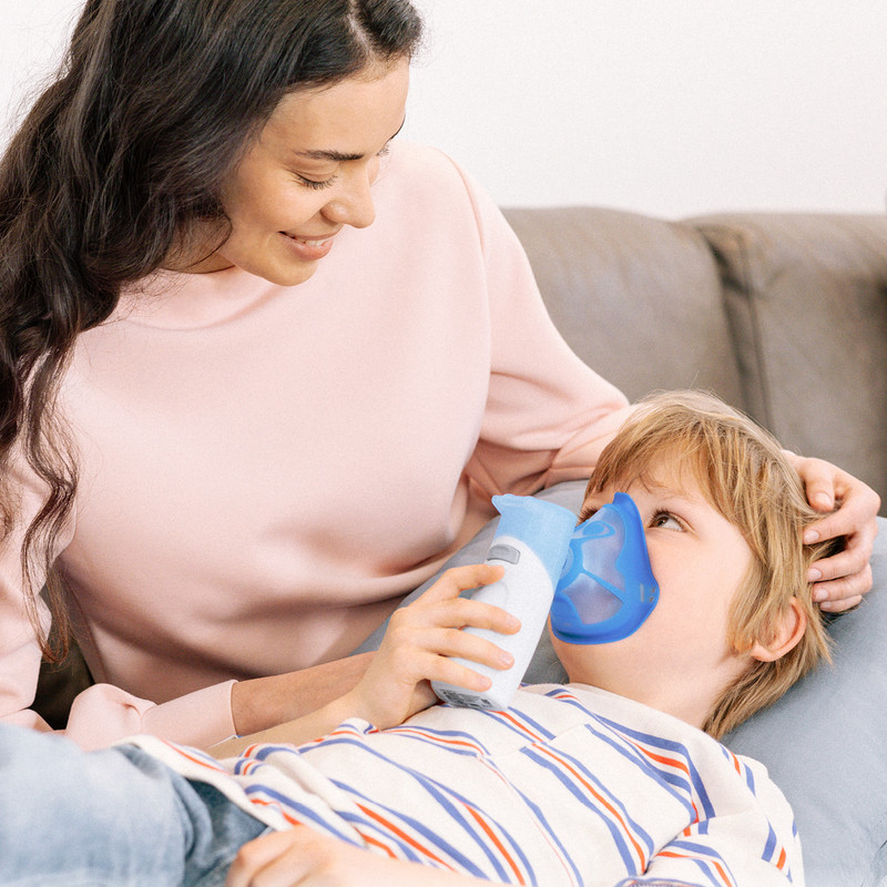 Kind liegt auf einem Sofa und inhaliert mit einem PARI BOY Go Inhalationsgerät mit Maske, betreuende Person sitzt daneben.