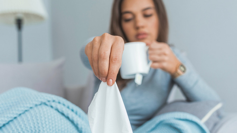 Woman sitting in bed under a blue blanket, holding a tissue and a white mug, appearing unwell or sick.
