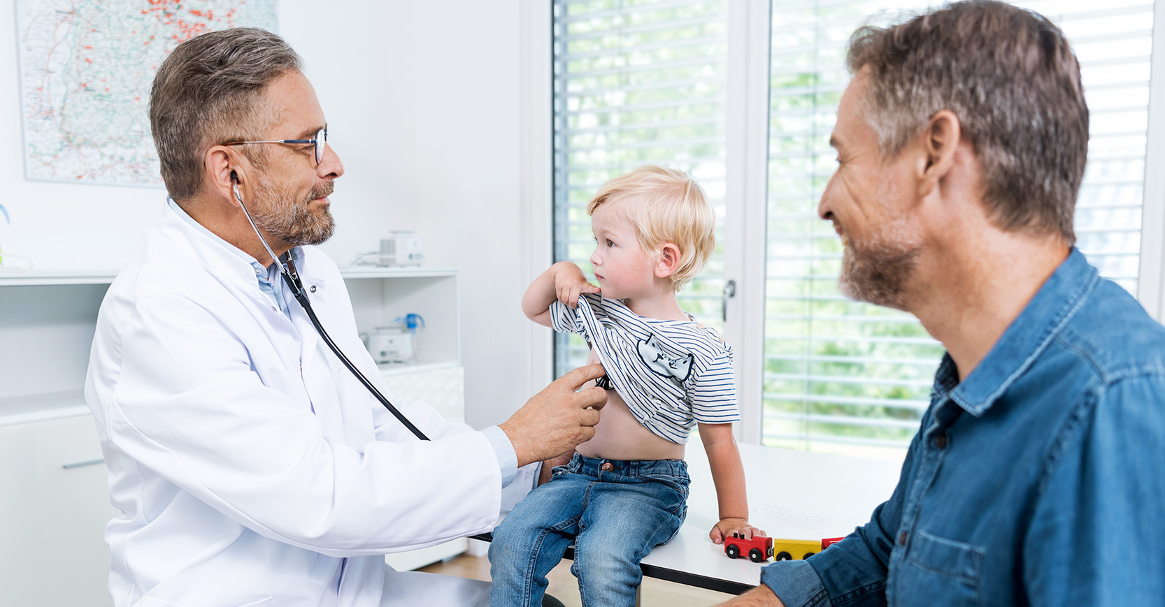 Ein Arzt hört einen kleinen Jungen an der Brust ab, sein Vater sitzt daneben