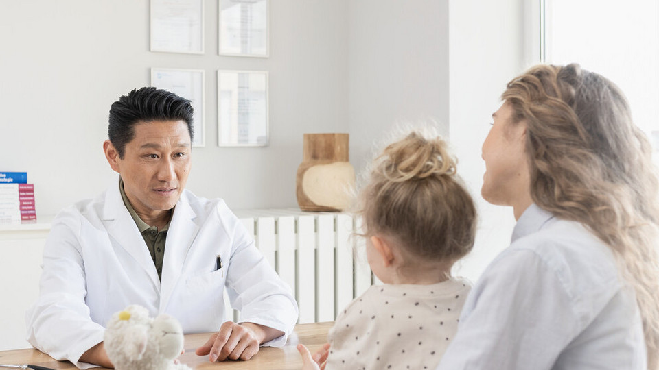 What is asthma? A simple explanation for children Junge Mutter sitzt mit Tochter auf dem Schoß dem Kinderarzt am Schreibtisch gegenüber