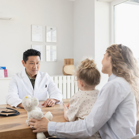 Junge Mutter sitzt mit Tochter auf dem Schoß dem Kinderarzt am Schreibtisch gegenüber