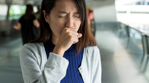 Hustenreiz stoppen: 8 Tipps, was Sie gegen einen Hustenanfall tun können Junge Frau hält sich an öffentlichem Ort beim Husten die Hand vor den Mund