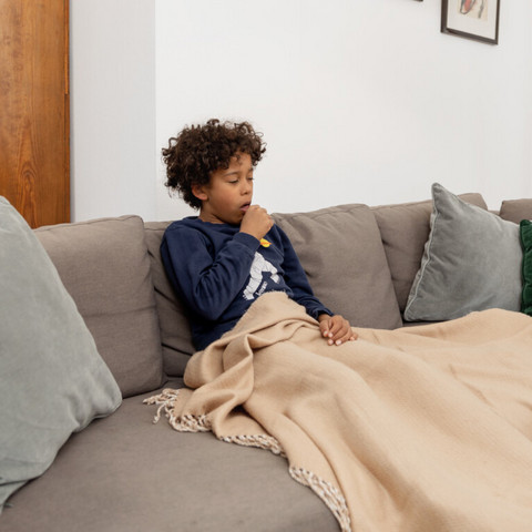 Junge sitzt auf Couch mit Decke und hustet in die Faust