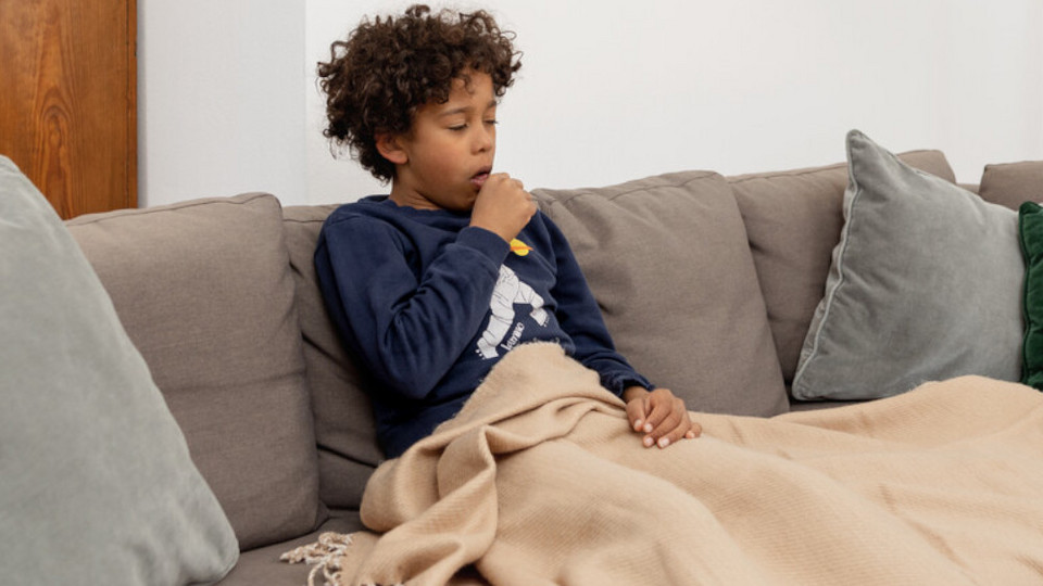 Junge sitzt auf Couch mit Decke und hustet in die Faust