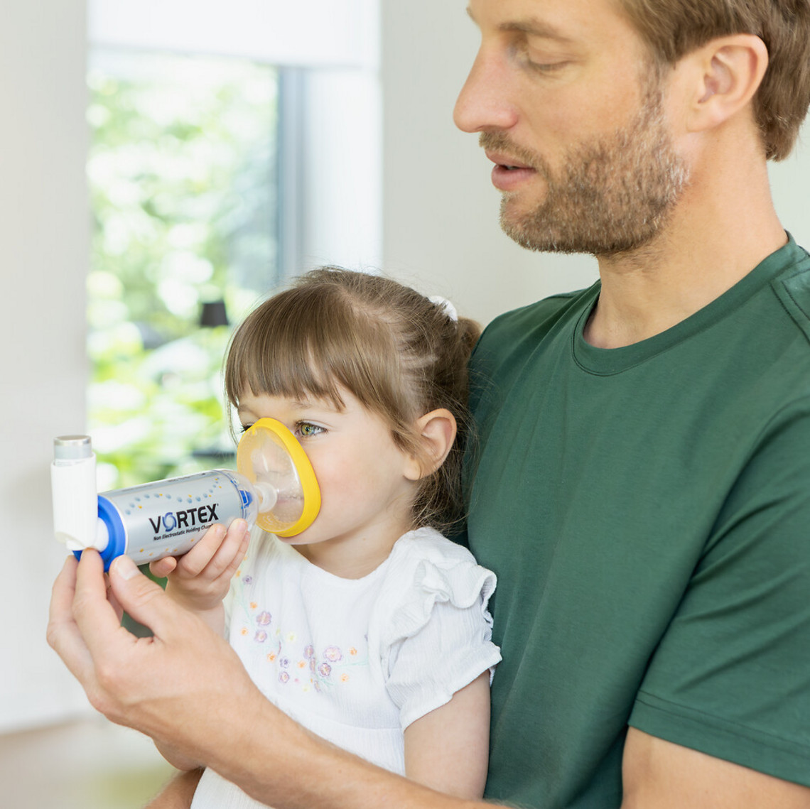 Vater hat Tochter auf dem Schoß und hilft ihr beim Inhalieren des Asthmaspray mit VORTEX Inhalierhilfe