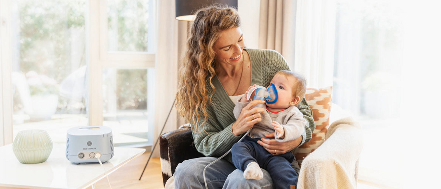 Eine Frau hält ihr Baby auf dem Schoß und hilft ihm, eine Inhalationsmaske zu benutzen. Im Hintergrund steht ein PARI COMPACT2 Inhalationsgerät.