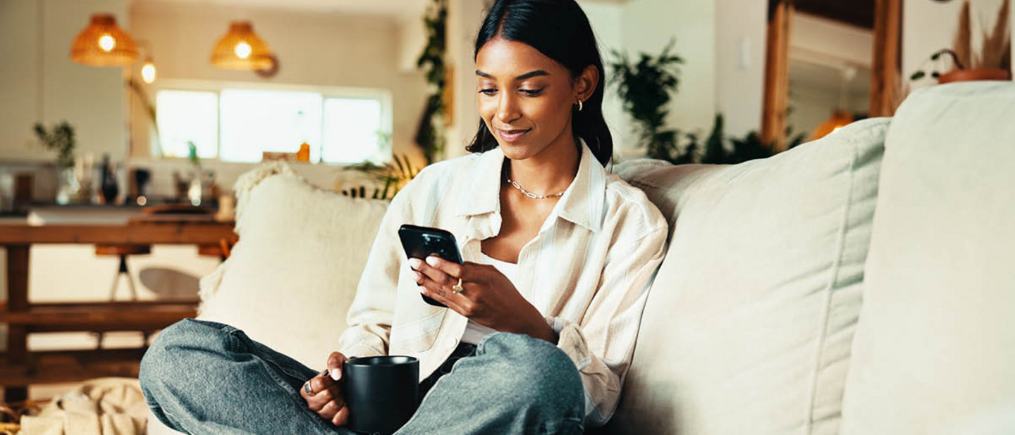 A person sits relaxed on the sofa, holding a smartphone and a cup in the living room.