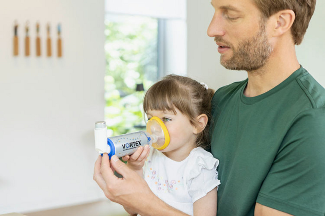 FAQ zu Inhalation bei Asthma: VORTEX Inhalierhilfe Junges Mädchen sitzt auf dem Schoß ihres Vaters und hält mit ihm ein Asthmaspray mit Vorschaltkammer vor den Mund.