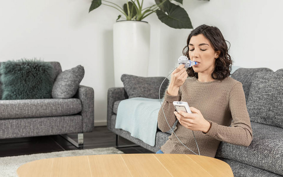 Mother seated on a sofa holding a young child on her lap while helping the child use a nebuliser connected to an inhalation device.